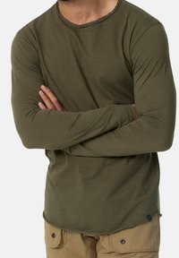 Olijfgroene shirt met lange mouwen en een ronde halslijn, met een gestikte zoom. De stof lijkt zacht en aansluitend. Draagt met beige cargobroek.