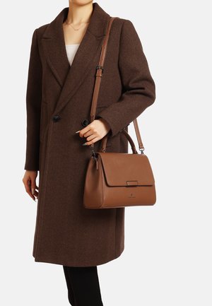 Cappotto a spina di pesce marrone, top chiaro, pantaloni neri, tiene in mano una borsa in pelle marrone con pattina e tracolla. Design strutturato e testurizzato.