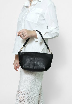 Donna con camicia bianca e gonna in pizzo che tiene in mano destra una borsa nera in pelle con trama e manico corto.