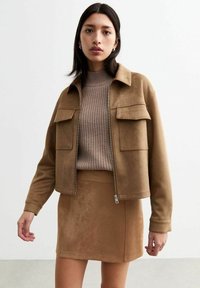Braune Wildleder-Zip-Jacke mit zwei vorderen Taschen, kombiniert mit einem passenden braunen Wildleder-Minirock und einem gerippten braunen Pullover darunter.