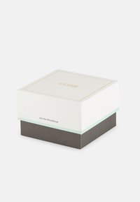Boîte CLUSE avec un dessus blanc portant des lettres dorées, une bande d'accent vert menthe et une base texturée gris foncé. Forme carrée avec couvercle.