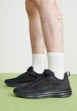Benen met witte geribbelde sokken en zwarte sportschoenen met getextureerde zolen, staand op een groene vloer tegen een effen muur.