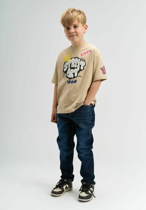 Beige graphic t-shirt met verschillende kleurrijke prints; donkerblauwe jeans met een rechte pasvorm; zwart-witte sneakers maken de outfit compleet.