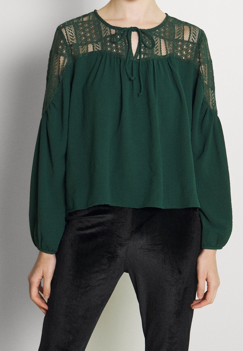 Femme portant un chemisier vert foncé avec des détails en dentelle aux épaules et des manches longues, associé à un pantalon en velours noir.
