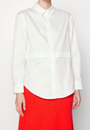 Chemise blanche à col, dotée d'une bande horizontale au niveau du ventre. Manches longues, texture lisse, ourlet rond.