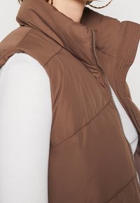 Brun pufferväst med hög krage, dragkedjestängning och quiltat mönster. Bärs över en vit långärmad topp, vilket visar en lagerdesign.