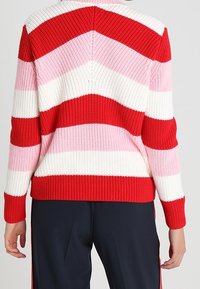 Pull en maille rayé rouge, rose et blanc avec un motif en chevrons. Présente une texture côtelée et un col arrondi, ajusté à la taille.