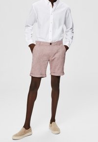 Une chemise blanche à boutons avec une texture subtile, associée à un short en lin rose clair et des chaussures beiges à enfiler. Une tenue propre et décontractée.