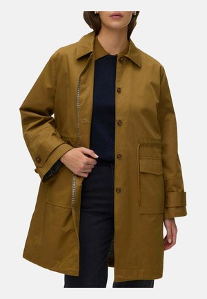 Femme portant un manteau beige boutonné avec de grandes poches avant sur un pull et un pantalon foncés, tenant le manteau légèrement ouvert d’une main.