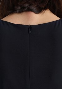 Robe noire en tissu lisse, avec une couture au dos et une fermeture éclair discrète au niveau du col. Design simple sans motifs ni accents.