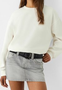 Sweatshirt blanc avec un col rond, associé à une jupe en denim gris clair. Une ceinture noire à clous se trouve à la taille, avec une boucle en argent.