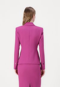Blazer rose ajusté à manches longues, avec des revers marqués et des accents en métal doré au niveau des poignets. Tissu lisse et structuré avec une silhouette cintrée.