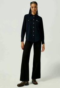Camisa de botão de manga longa azul-escura com dois bolsos no peito, combinada com calças largas pretas e botas de tornozelo castanhas. Tecido simples, design casual.
