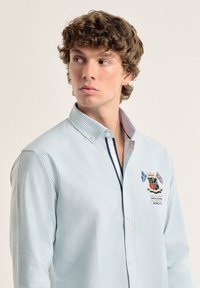 Camisa de botones de rayas azul claro con cuello, que presenta un logo bordado con detalles. Tela suave, mangas largas y un corte ajustado.