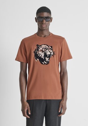 Jonge man met donkere zonnebril en een roestkleurig T-shirt met een grote grafische afbeelding van een brullende tijger aan de voorkant, staand tegen een effen achtergrond.