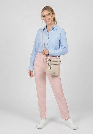 Junge Frau lächelt, trägt ein hellblau kariertes Hemd, pinke Hosen, weiße Sneakers und hält eine beige Umhängetasche mit vorderen Reißverschlüssen.