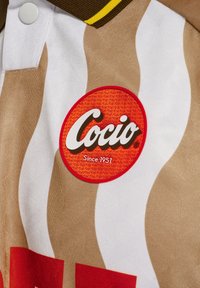 Brun- och vitrandig kortärmad polo med en röd cirkulär "Coccio" logotypapplikation med vit text, strukturväv, och gula krageaccenter.