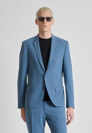 Slim-fit lichtblauw pak met zwart overhemd eronder, gedragen door een persoon met kort blond haar en donkere zonnebril, staand tegen een effen grijze achtergrond.