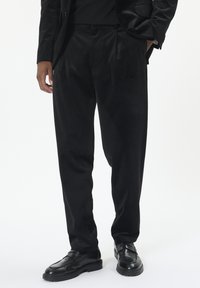 Homme portant un pantalon noir ample, un blazer noir et des mocassins en cuir noirs, se tenant avec une main dans la poche contre un fond blanc.