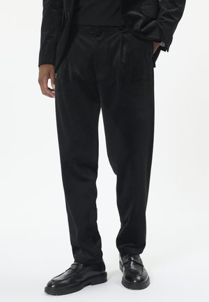 Mann trägt lockere schwarze Hose, schwarze Blazer und schwarze Lederloafers, steht mit einer Hand in der Tasche vor weißem Hintergrund.