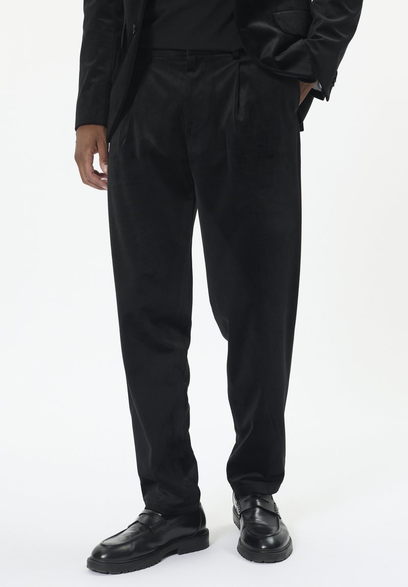 Homme portant un pantalon noir ample, un blazer noir et des mocassins en cuir noirs, se tenant avec une main dans la poche contre un fond blanc.