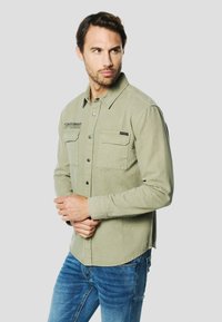 Chemise boutonnée vert olive en coton doux avec deux poches poitrine, boutons-pression, et un ourlet arrondi, portée avec un jean denim bleu.