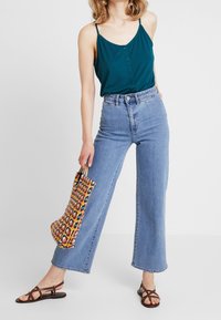 Tealfärgad ärmlös topp med knappar, högmidjade blå jeans och en flerfärgad mönstrad tote-väska. Bruna remsandaler fulländar looken.