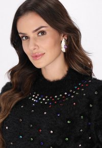 Sort fuzzy sweater med farverige rhinestones arrangeret i et mønster. Modellen bærer store guldfarvede øreringe formet som blade med lilla detaljer.