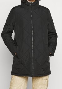 Svart quiltad jacka med hög krage, framdragkedja och två sidofickor. Exteriören har en slät yta med diamantmönstrad söm.