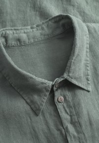 Camicia di lino verde con colletto alla francese e due bottoni marroni, realizzata in un tessuto testurizzato e leggermente stropicciato.