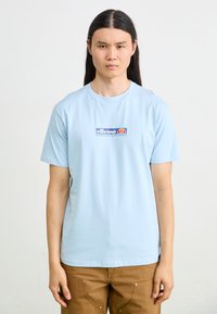 Light blue cotton t-shirt with short sleeves, featuring a graphic logo in red and blue on the chest. Plain design, crew neckline.