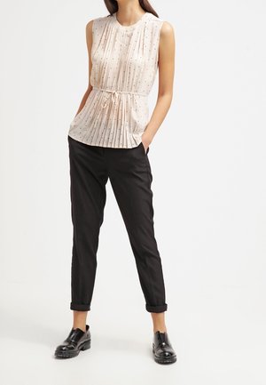 Vrouw draagt een mouwloze crèmekleurige blouse met zwarte polka dots en plooien, zwarte broek met omslag en zwarte leren loafers, staand met handen in de zakken.