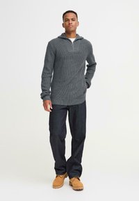 Grå strikket genser med glidelås krage, kombinert med mørke denimjeans og brune støvler. Teksturert stoff og avslappet passform.