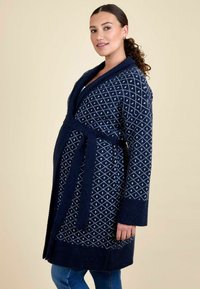 Femeie cu părul strâns la spate, îmbrăcată într-un cardigans din tricot cu model, de culoare albastru închis, având o curea, stând în fața unui fundal bej.