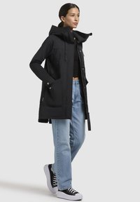 Veste noire imperméable avec un col montant, capuche ajustable, deux poches avant et une coupe droite, accompagnée d'un jean bleu clair et de baskets noires.