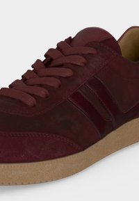 Baskets en daim rouge foncé avec lacets assortis, deux bandes latérales rouge foncé et une semelle en caoutchouc brun clair texturée.
