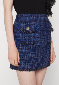 Mini-jupe en tweed bleu avec un ourlet frangé, deux poches avant avec des boutons dorés, et un motif texturé de lignes bleues plus foncées et plus claires.