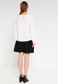 Blusa bianca a maniche lunghe con dettagli in pizzo e scollatura a chiave sul retro, abbinata a una gonna nera. Il modello indossa scarpe con tacco quadrato nere.