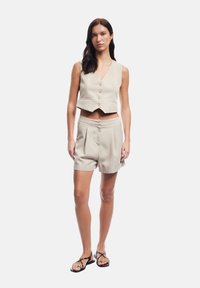 Canotta corta beige chiaro con chiusure a bottone e pantaloni a shorts coordinati a vita alta. L'outfit presenta un design minimalista e una trama di tessuto liscia.