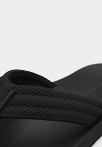 Μαύρο σανδάλι με υφή και ρυθμιζόμενο λουρί, με μαξιλαρωτό πέλμα και μικρή ετικέτα με μερική σήμανση στη πλευρά του λουριού.