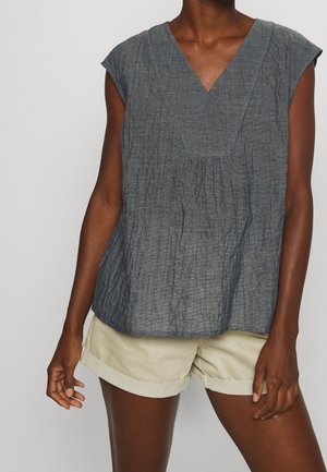 Personne portant un haut sans manches gris foncé texturé à col en V et un short beige clair avec ourlet roulé, debout devant un fond uni.