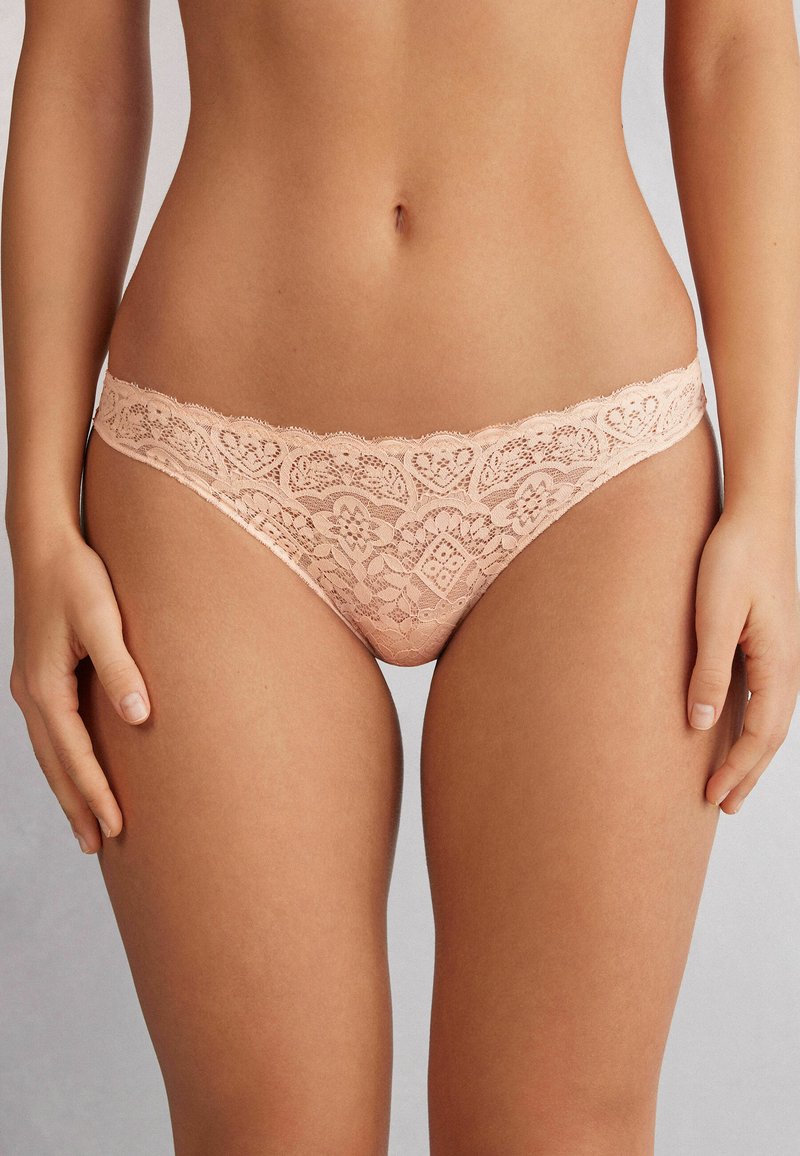 String en dentelle couleur pêche douce avec des motifs floraux complexes, des bords ondulés et une finition délicate autour de la taille.