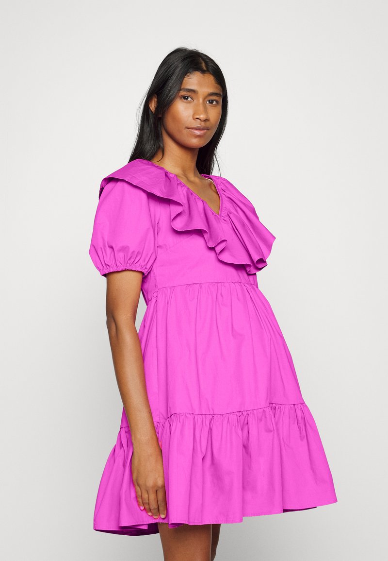 Vestido de algodão fúcsia com decote assimétrico, mangas curtas e bufantes, e saia rodada com bainha em camadas. Textura suave e cor vibrante.