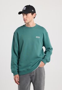 Teal sweatshirt with round neckline and NYC logo, paired with a black cap. Model wears gray jeans, showing a casual layered look.