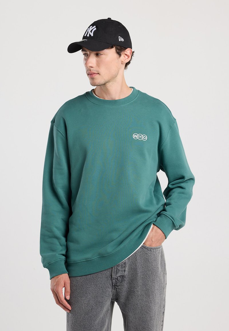 Teal sweatshirt with round neckline and NYC logo, paired with a black cap. Model wears gray jeans, showing a casual layered look.