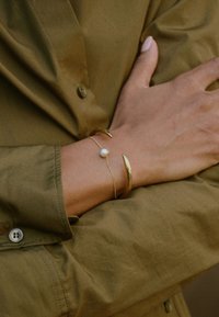 Gold-tone metal bracelets on a wrist; one features a central pearl, while the other has a curved, elongated design. Olive green shirt visible.