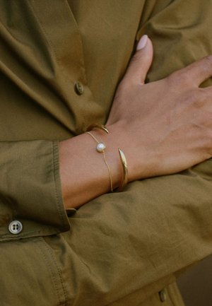 Gold-tone metal bracelets on a wrist; one features a central pearl, while the other has a curved, elongated design. Olive green shirt visible.