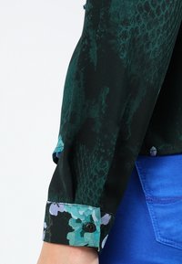 Blouse à manches longues en vert foncé avec un motif serpent. Les poignets présentent un imprimé floral et un bouton noir. Portée avec un bas en tissu bleu.
