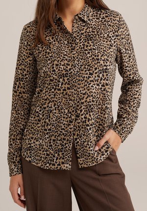 Femme portant une chemise à manches longues à boutons imprimée léopard marron et noire, avec un pantalon marron, main dans la poche, fond neutre.