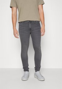 Jeans ajustados grises con cinco bolsillos, material de denim y un acabado suave, combinados con una camiseta beige y zapatillas gris claro.